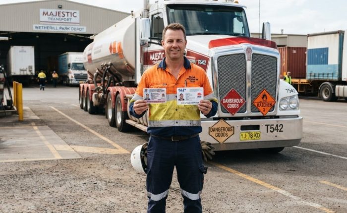 NSW truck driver in high-vis vest holding dangerous goods licence certificate and SLP Safe Load Program card at a transport depot in Wetherill Park NSW