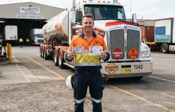 NSW truck driver in high-vis vest holding dangerous goods licence certificate and SLP Safe Load Program card at a transport depot in Wetherill Park NSW